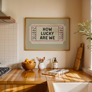 May include: A framed print featuring a ticket design with the text "HOW LUCKY ARE WE" in black and pink. The print is displayed above a wooden countertop with a bowl of oranges, utensils, and a soap dispenser.