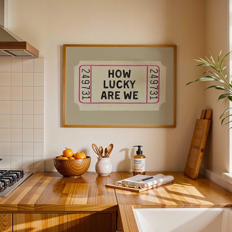 May include: A framed print featuring a ticket design with the text "HOW LUCKY ARE WE" in black and pink. The print is displayed above a wooden countertop with a bowl of oranges, utensils, and a soap dispenser.
