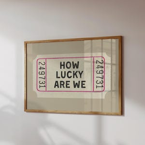 May include: A framed print featuring a ticket design with the text "HOW LUCKY ARE WE" in black. The ticket has pink borders and the numbers "249731" on each end. The frame is made of wood.