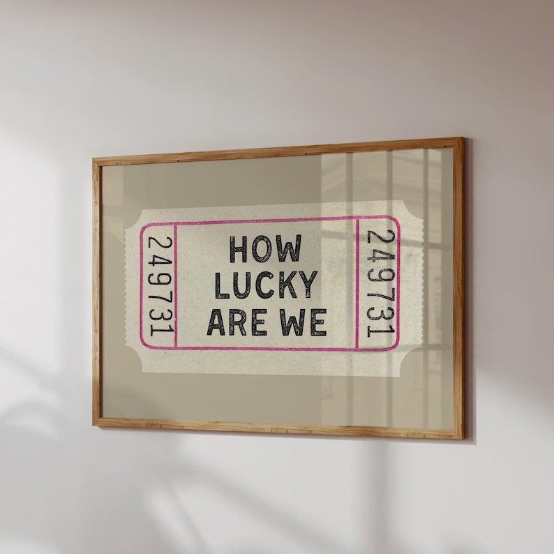 May include: A framed print featuring a ticket design with the text "HOW LUCKY ARE WE" in black. The ticket has pink borders and the numbers "249731" on each end. The frame is made of wood.