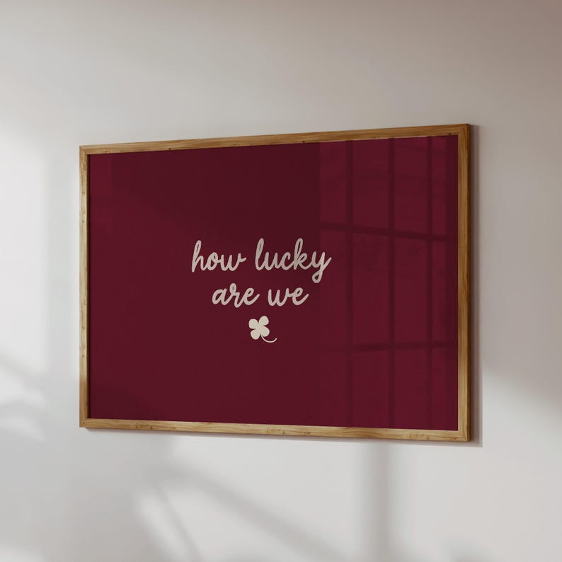 May include: A framed print with a deep red background and the words "how lucky are we" in a cursive font. A small four-leaf clover graphic is below the text. The frame is made of wood.