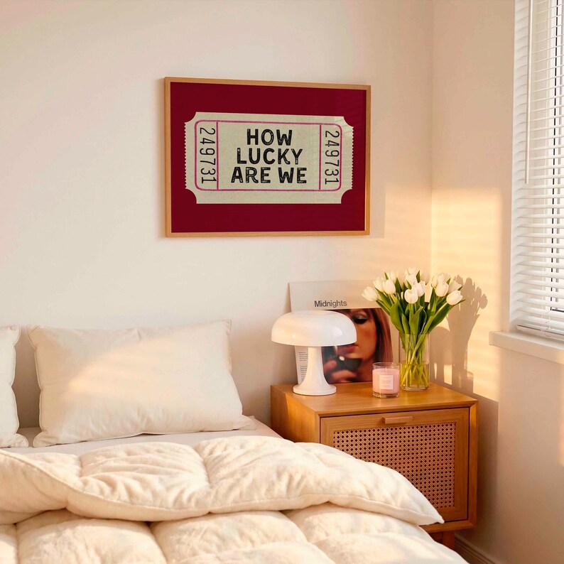 May include: A framed print with the text "HOW LUCKY ARE WE" in a ticket design. The print is in a red frame and hangs above a bed with white bedding. A nightstand with a lamp, flowers, and a candle is next to the bed.