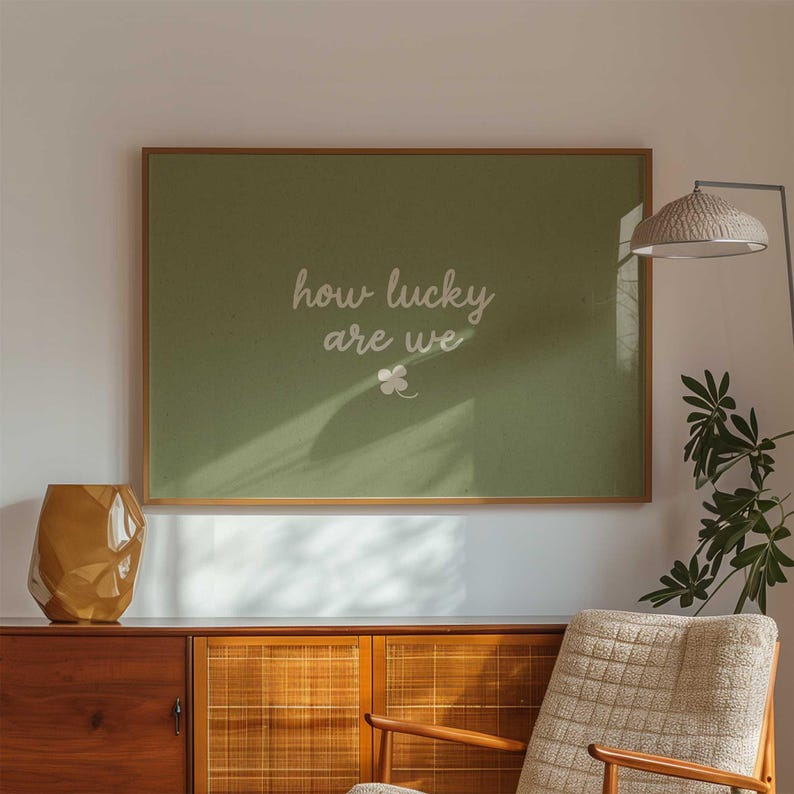 May include: A framed wall art piece with a green background and the text "how lucky are we" in white script. A small white clover graphic is below the text. The frame is a light brown color. The art is displayed above a wooden cabinet.