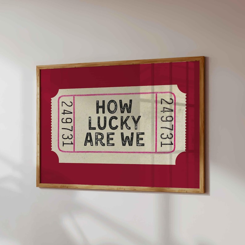 May include: A framed print featuring a vintage-style ticket design. The ticket is cream-colored with the text "HOW LUCKY ARE WE" in black and pink. The frame is a light brown color, and the background is a deep red.
