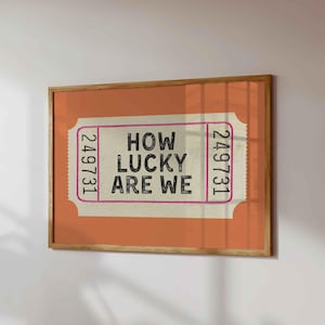 May include: A framed print featuring a vintage-style ticket design. The ticket has the text "HOW LUCKY ARE WE" in black, with the number "249731" on each side. The background is orange, and the frame is wood.