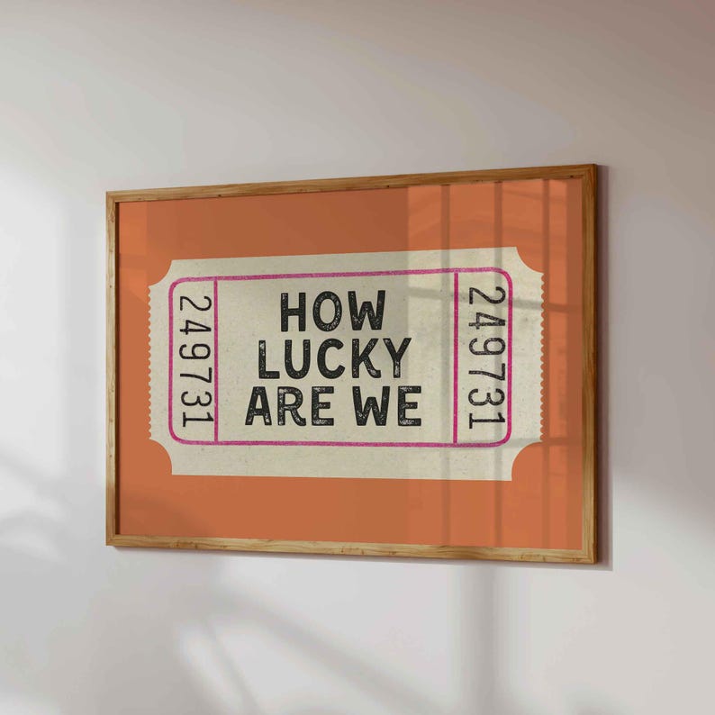 May include: A framed print featuring a vintage-style ticket design. The ticket has the text "HOW LUCKY ARE WE" in black, with the number "249731" on each side. The background is orange, and the frame is wood.