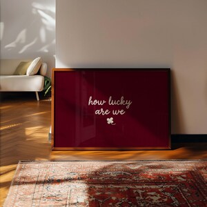 May include: A framed print with a deep red background and the words "how lucky are we" in white script. A small white four-leaf clover is below the text. The frame is a warm brown color.