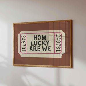 May include: A framed print featuring a ticket design with the text "HOW LUCKY ARE WE" in black. The ticket has a pink border and the number "249731" on each end. The frame is a warm brown color.