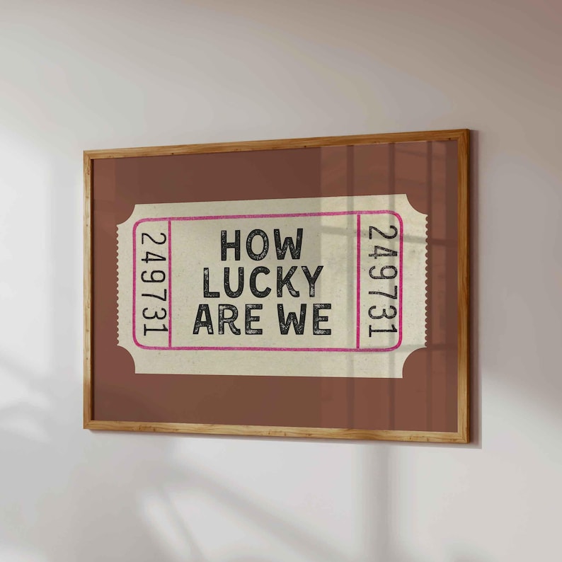 May include: A framed print featuring a ticket design with the text "HOW LUCKY ARE WE" in black. The ticket has a pink border and the number "249731" on each end. The frame is a warm brown color.