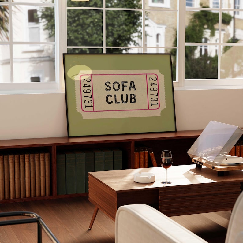 May include: A framed print with the text "SOFA CLUB" in a ticket design. The print is displayed on a wooden cabinet with books and a record player. A glass of red wine and an ashtray are on a wooden coffee table.