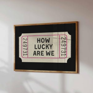 May include: A framed print featuring a vintage-style ticket design. The ticket has the text "HOW LUCKY ARE WE" in black, with the numbers "249731" in pink on each side. The frame is a natural wood color.