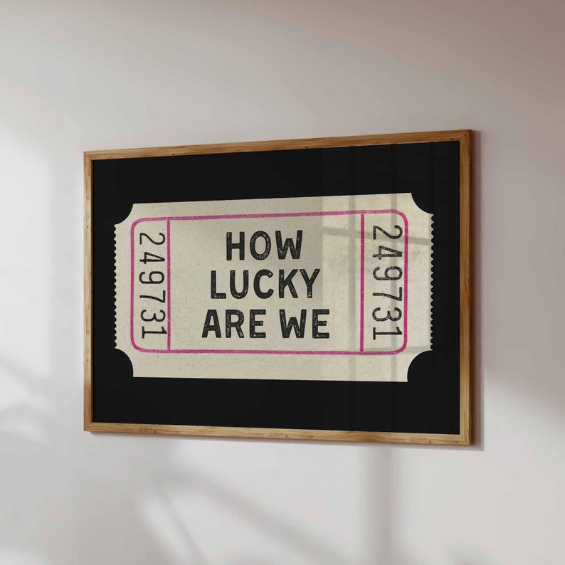 May include: A framed print featuring a vintage-style ticket design. The ticket has the text "HOW LUCKY ARE WE" in black, with the numbers "249731" in pink on each side. The frame is a natural wood color.