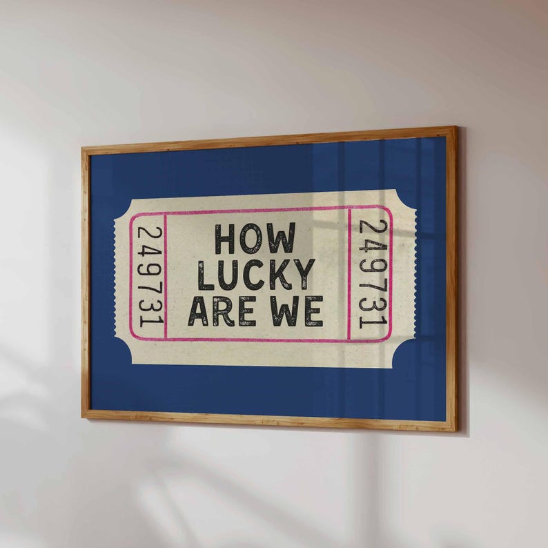 May include: A framed print featuring a ticket design with the text "HOW LUCKY ARE WE". The ticket has a cream background with pink borders and the number "249731" on each side. The frame is a natural wood color, and the background is a deep blue.