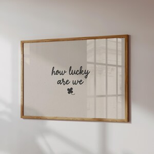 'How Lucky Are We' print. Available in 8 different colour options. The poster in the picture has a brown background with the words 'how lucky are we' in small cursive beige font in the centre, with a small 4 leaf clover shamrock just beneath it. Displayed in a large horizontal wooden frame on a neutral wall.
