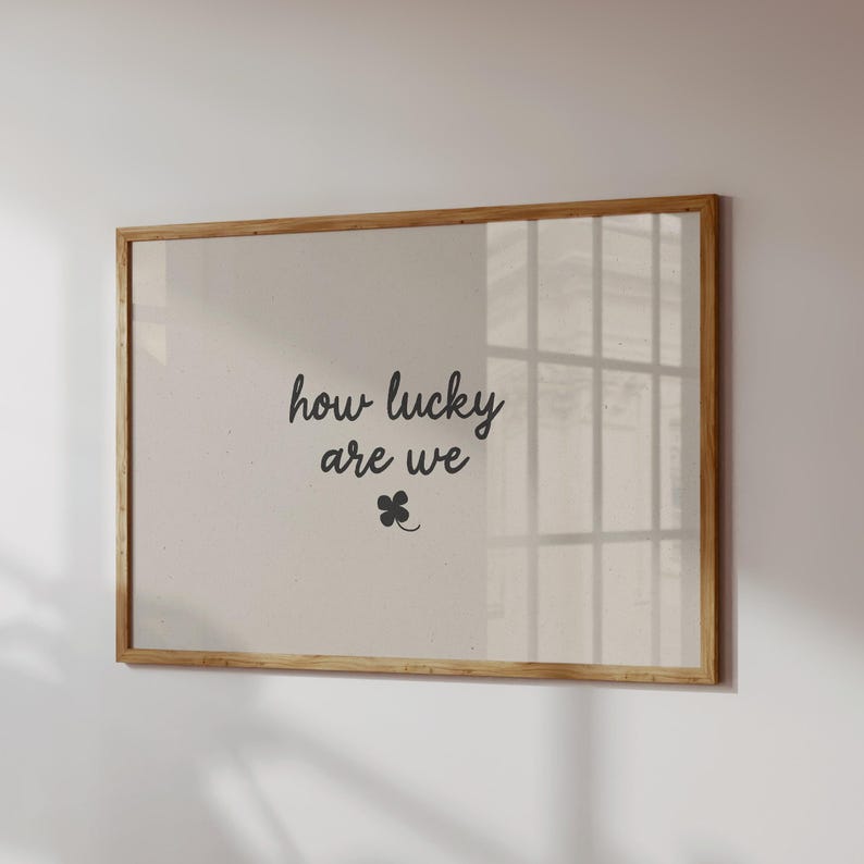 'How Lucky Are We' print. Available in 8 different colour options. The poster in the picture has a brown background with the words 'how lucky are we' in small cursive beige font in the centre, with a small 4 leaf clover shamrock just beneath it. Displayed in a large horizontal wooden frame on a neutral wall.
