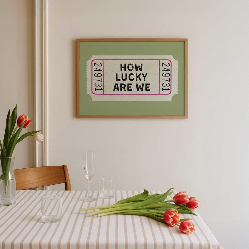 May include: A framed print featuring a ticket design with the text "HOW LUCKY ARE WE" in black. The print is set against a light green background within a wooden frame. The image is displayed above a table set with glasses, tulips, and a striped tablecloth.