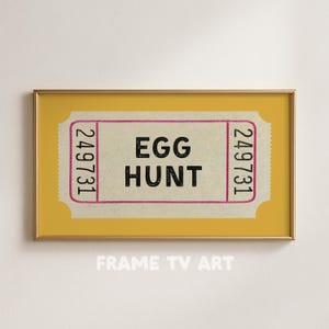 May include: A framed print featuring a vintage-style ticket with the words "EGG HUNT" in bold black letters. The ticket has a light beige background with pink borders and the number "249731" on each side. The frame is gold.