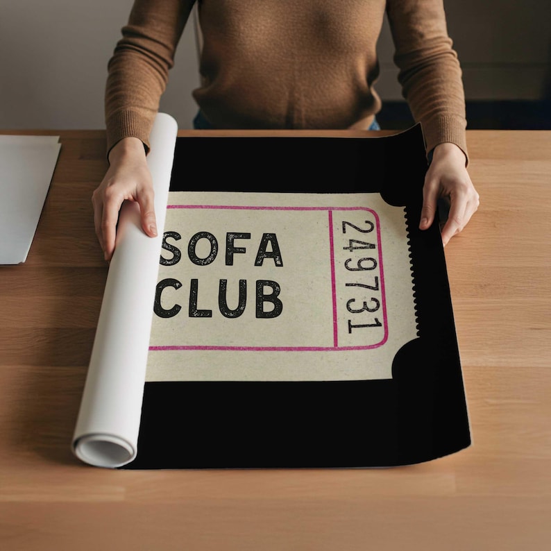 May include: A person unrolling a black poster with a vintage ticket design. The poster features the words "SOFA CLUB" in a bold, black font, with a pink border and the number "249731". The poster is on a wooden surface.