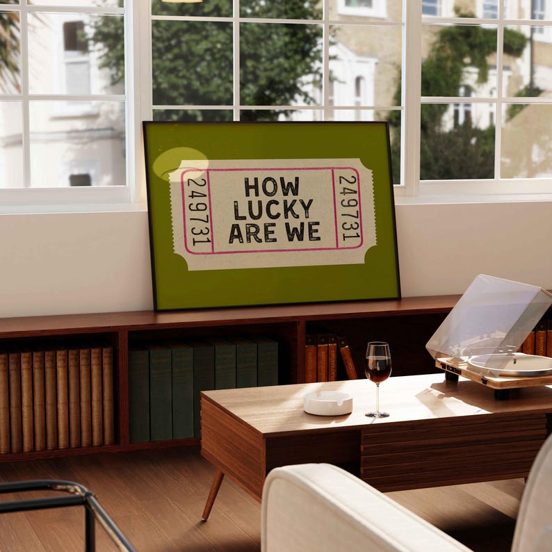 May include: A framed print with the text "HOW LUCKY ARE WE" on a ticket design. The print is set against a green background and framed in black. The image is displayed on a wooden cabinet in a room with a window.