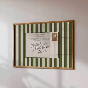 May include: A rectangular wall art piece in a wooden frame featuring a vertical dark green and white striped background. Centered is a postcard graphic with the text "POST CARD" and the cursive script "It feels so good to be home."