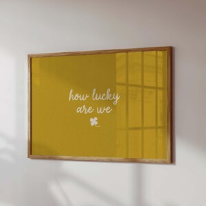 May include: A framed print with a mustard-yellow background. The white cursive text reads "how lucky are we" with a small four-leaf clover below. The frame is made of light-colored wood.