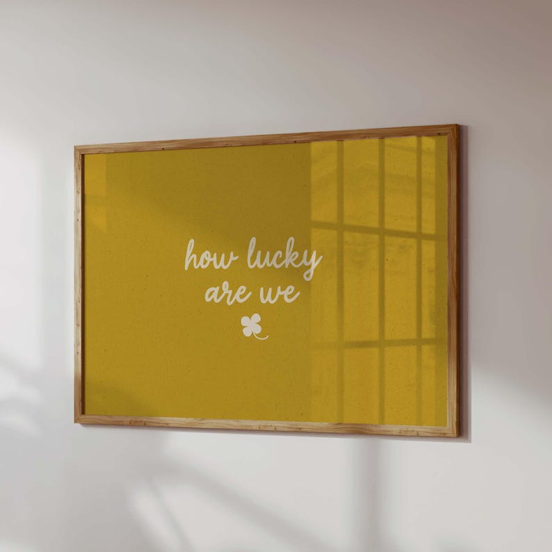 May include: A framed print with a mustard-yellow background. The white cursive text reads "how lucky are we" with a small four-leaf clover below. The frame is made of light-colored wood.