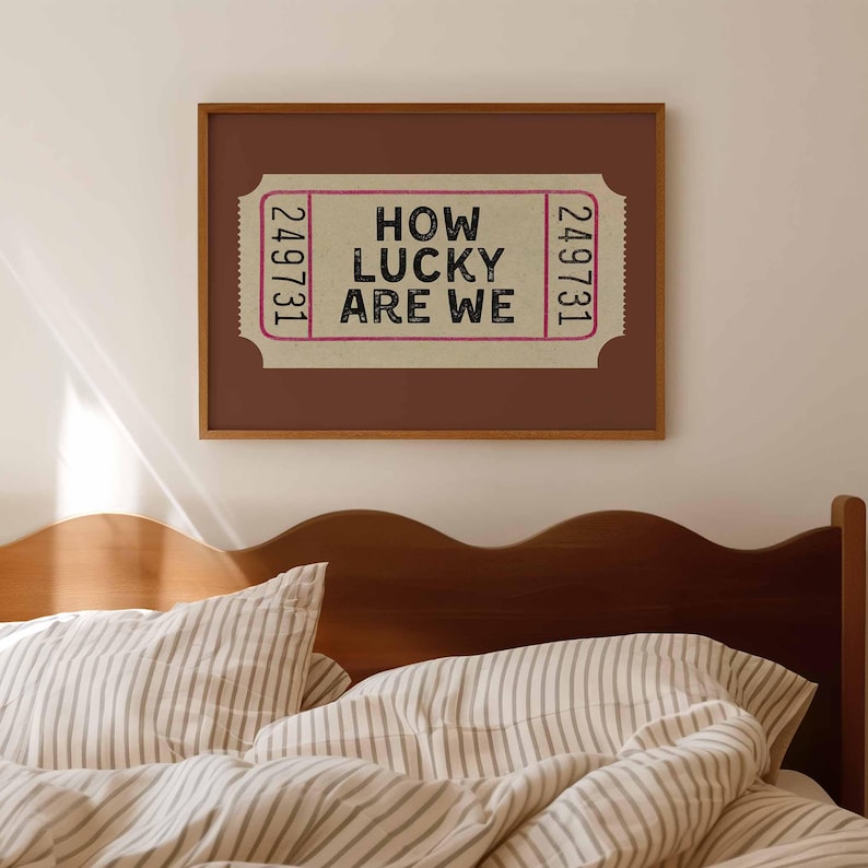 May include: A framed print featuring a vintage-style ticket design with the text "HOW LUCKY ARE WE" in black. The ticket is set against a light pink background, framed in a brown wooden frame. The print is displayed above a bed with striped bedding.