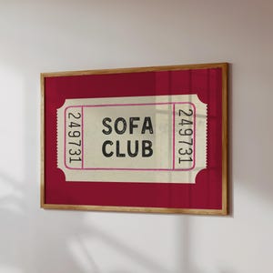 May include: A framed print featuring a vintage-style ticket design. The ticket, set against a deep red background, reads "SOFA CLUB" in black text. The ticket has pink borders and the number "249731" on each side. The frame is made of wood.