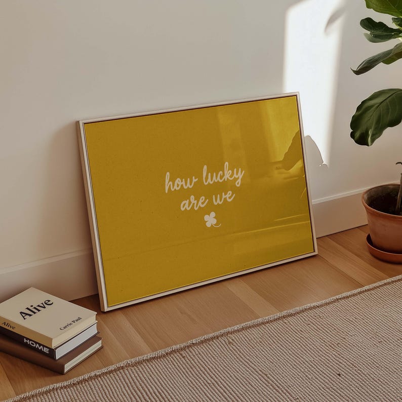 May include: A framed yellow print with the words "how lucky are we" in white script, along with a small clover graphic. The frame is white. The print is leaning against a white wall. Books are stacked on the floor.