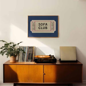 May include: A framed print featuring a ticket design with the words "SOFA CLUB" in a retro font. The print is set against a navy blue background and framed in a wooden frame. A vintage record player and vinyl records are on a wooden cabinet.