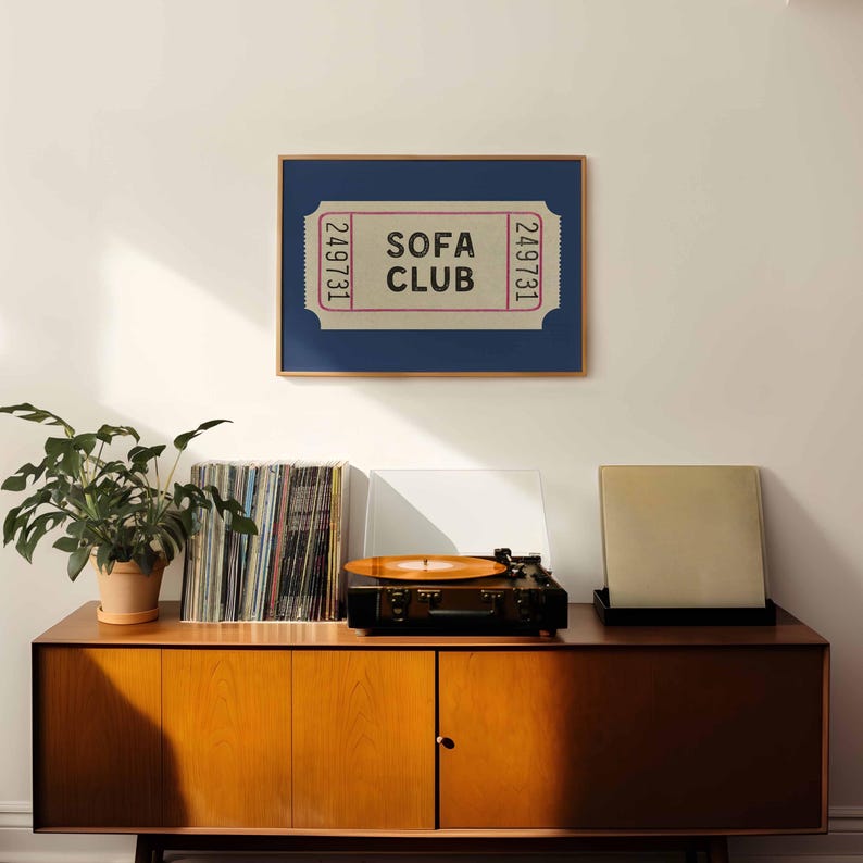 May include: A framed print featuring a ticket design with the words "SOFA CLUB" in a retro font. The print is set against a navy blue background and framed in a wooden frame. A vintage record player and vinyl records are on a wooden cabinet.