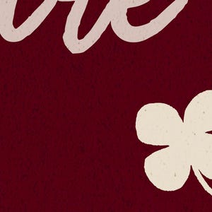 May include: A burgundy background with a textured appearance. The upper left corner features the word "love" in a cream-colored, cursive font. A cream-colored four-leaf clover is in the lower right corner.