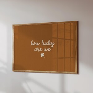 May include: A framed wall art piece with a warm brown background. The white cursive text reads "how lucky are we" with a small four-leaf clover below. The frame is made of light-colored wood.