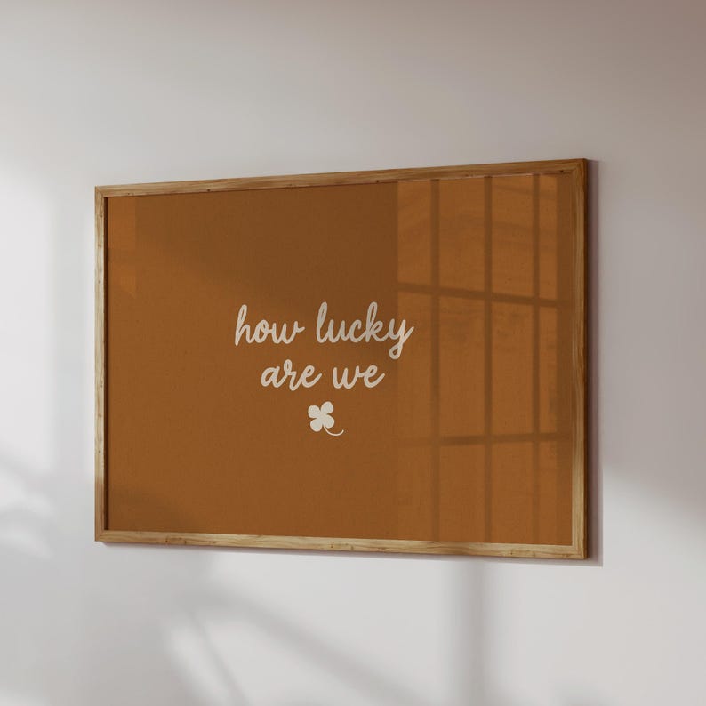 May include: A framed wall art piece with a warm brown background. The white cursive text reads "how lucky are we" with a small four-leaf clover below. The frame is made of light-colored wood.