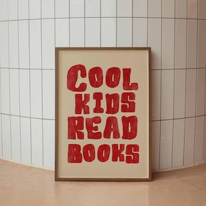 May include: A framed print featuring the phrase "COOL KIDS READ BOOKS" in red, bold letters. The text is set against a beige backdrop, within a brown wooden frame. The print is displayed against a white tiled wall and a pinkish-beige floor.