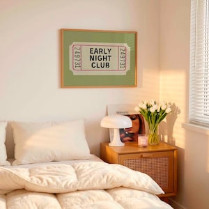 May include: A bedroom interior with a framed print reading "EARLY NIGHT CLUB." A bedside table holds a lamp, a vase of white tulips, and a candle. A bed with white bedding is in the foreground, with a window to the right.