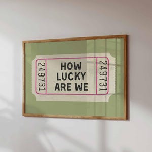 May include: A framed print featuring a ticket design with the text "HOW LUCKY ARE WE" in black. The ticket is set against a light green background, framed in a natural wood tone.