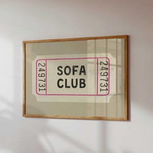 May include: A framed print featuring a vintage-style ticket design. The ticket has the words "SOFA CLUB" in black text, framed by a pink border, and the number "249731" on each end. The frame is made of light brown wood.