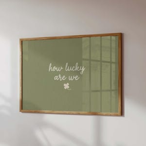May include: A framed print with a light brown wooden frame. The print has a sage green background with the words "how lucky are we" in white script, and a small white four-leaf clover.