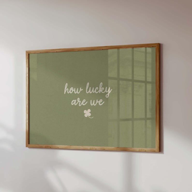 May include: A framed print with a light brown wooden frame. The print has a sage green background with the words "how lucky are we" in white script, and a small white four-leaf clover.