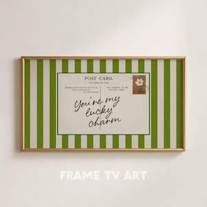 Puede incluir: Una impresión enmarcada con un diseño de postal vintage. La postal tiene el texto "You're my lucky charm" en cursiva, un sello floral y un fondo a rayas verdes y blancas. El marco es dorado.