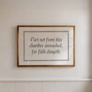 Funny printable bathroom wall art that says 'part not from this chamber unwashed, for filth clingeth' in black medieval font with medieval style border on a beige background. Displayed in a horizontal wooden frame hanging on a neutral wall.
