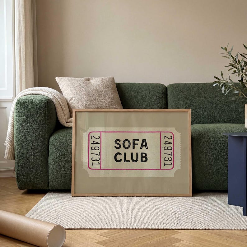 May include: A framed print featuring the text "SOFA CLUB" in black, set against a light beige background, resembling a ticket. The print is in a light wooden frame, placed in front of a green sofa. A beige rug and a rolled-up poster are in the foreground.