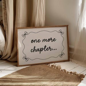Printable bookish wall art featuring the words 'one more chapter...' in a handwritten style font with a wavy border and a bow in each corner. Black illustration on a beige background. Displayed in a large horizontal wooden frame in a neutral room.