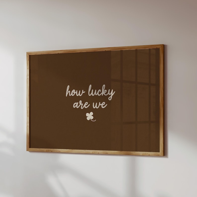 May include: A framed wall art piece with a brown background and a wooden frame. The text "how lucky are we" is written in white script, with a small four-leaf clover below the text.