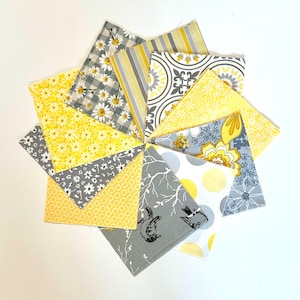 May include: A collection of fabric squares arranged in a circular pattern. The squares feature various patterns, including daisies, stripes, and floral designs, in shades of yellow, gray, and white. The arrangement creates a visually appealing, decorative display.