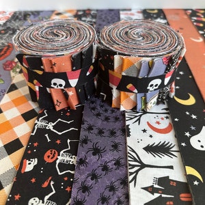 May include: Two rolled fabric bundles with Halloween-themed patterns. The bundles are wrapped with black fabric featuring candy corn and skull designs. Surrounding strips display skeletons, spiders, and haunted house motifs in orange, purple, and black.
