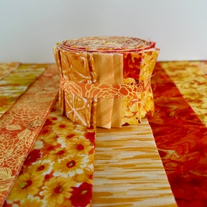 May include: A roll of fabric scraps in shades of orange, yellow, and red. The fabric is patterned with flowers, swirls, and stripes. The roll is sitting on a table with several strips of fabric laid out in front of it.