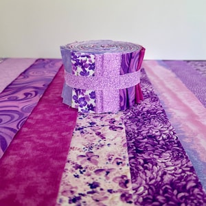 May include: A roll of purple fabric strips, featuring various patterns including floral and abstract designs. The fabric is held together with a matching purple band. The image showcases a variety of shades of purple.