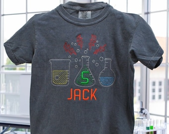 May include: A dark gray t-shirt with an embroidered design of beakers and lightning bolts. The text "JACK" is embroidered in orange below the beakers. The number "5" is embroidered in green inside one of the beakers.